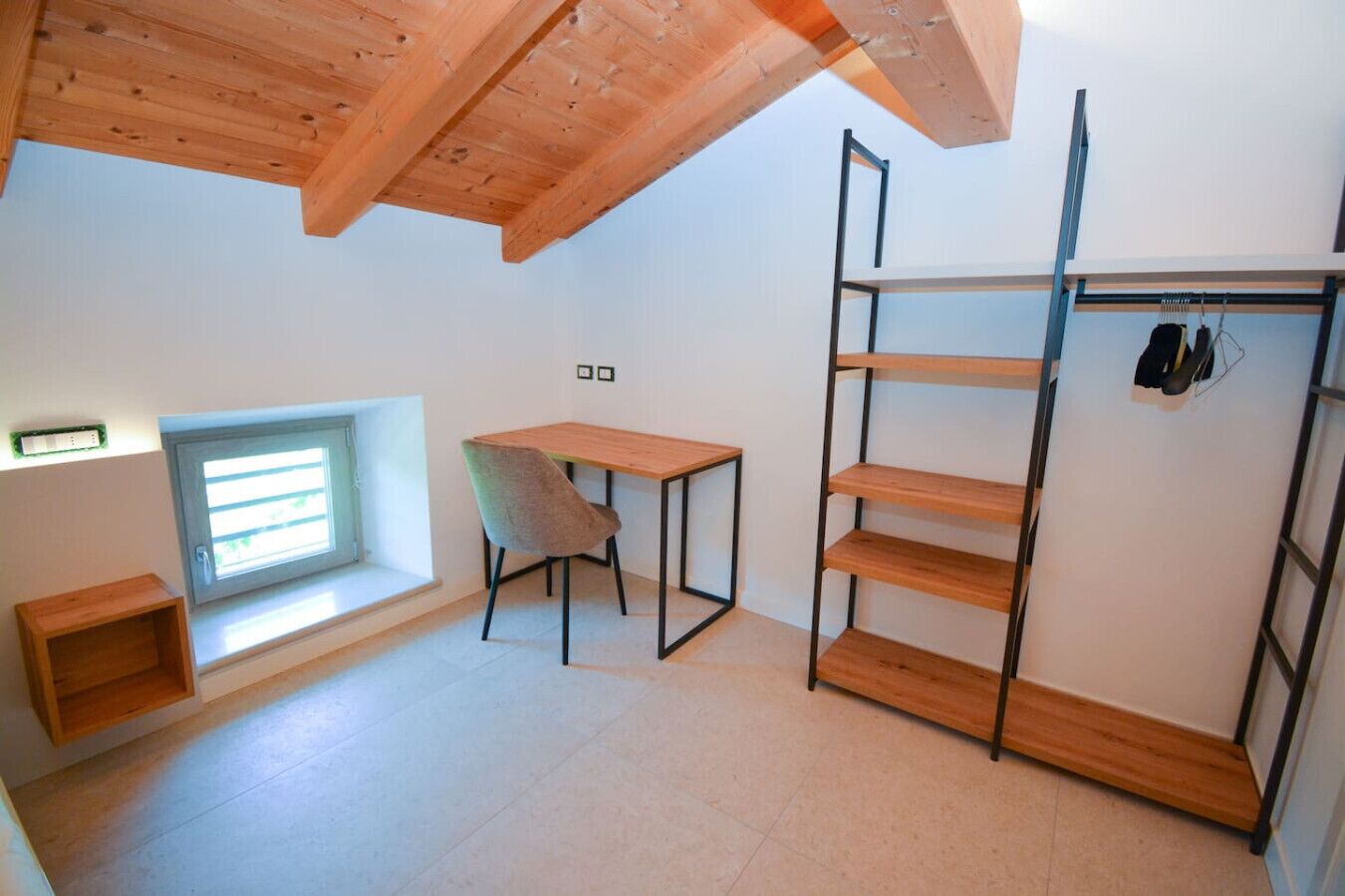 Una stanza con soffitto spiovente, un tavolino con una sedia, una finestra con persiane e una scaffalatura in metallo.