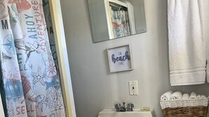 Un baño con cortina de ducha de motivos náuticos, un lavabo blanco, un espejo, una cita enmarcada, un inodoro y una cesta de toallas.