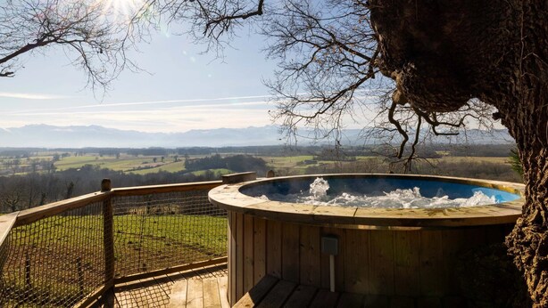 Un jacuzzi con cubierta de madera y baranda metálica, con vistas a un paisaje pintoresco con montañas al fondo.