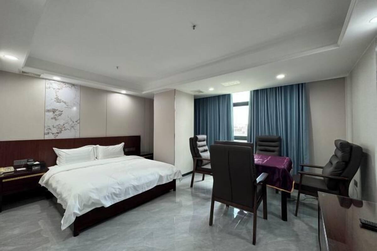 A hotel room with a bed, a desk, two chairs, a nightstand, and a window with curtains.