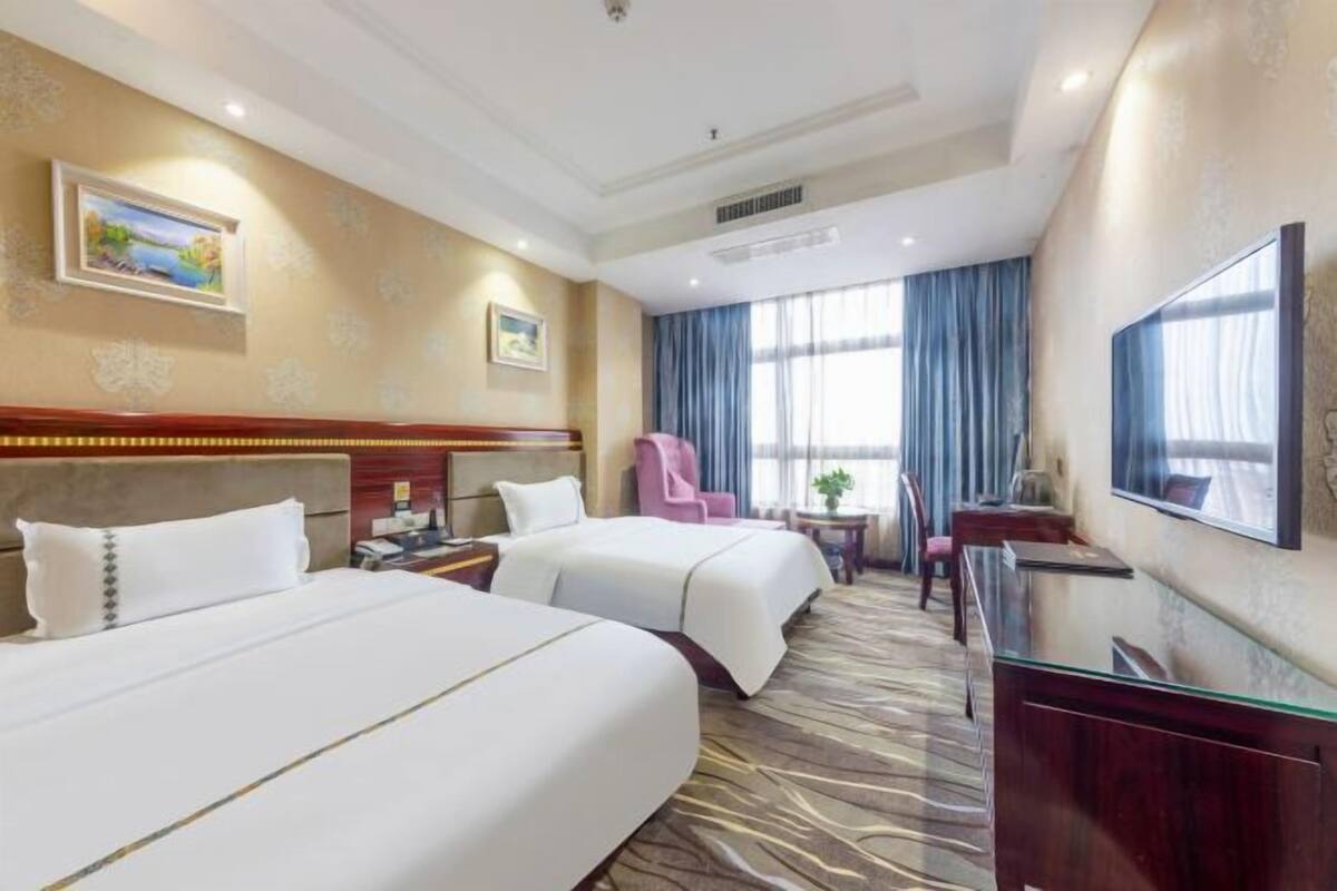 A hotel room with two beds, a desk, a chair, a TV, and a window with curtains.
