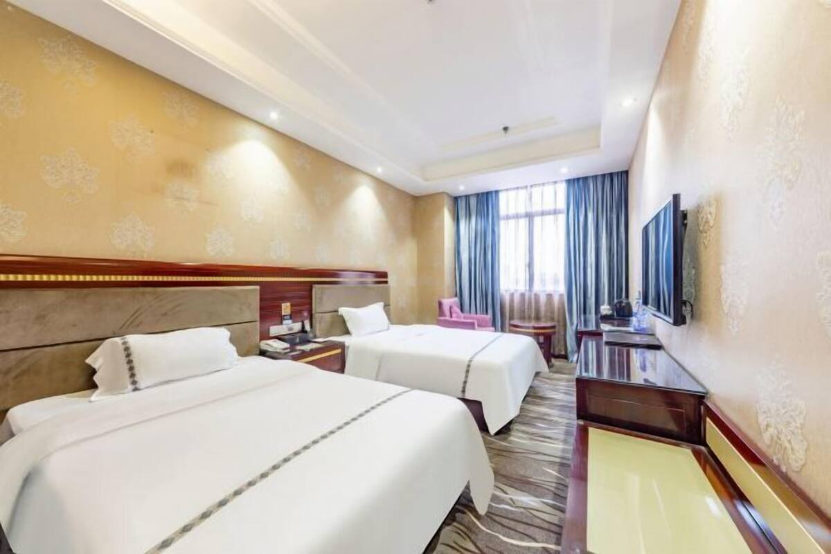 A hotel room with two beds, a desk, a chair, a TV, and a window with curtains.