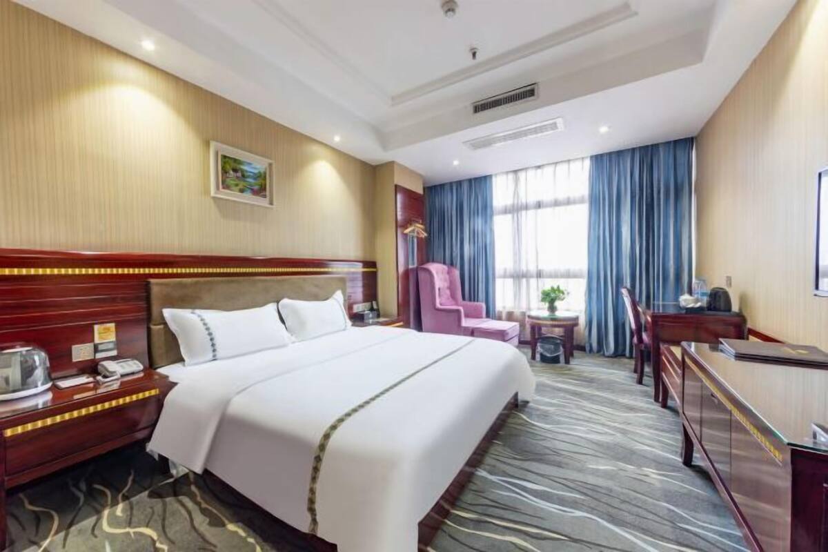 A hotel room with a large bed, a desk, a chair, a small table, and a window with curtains.