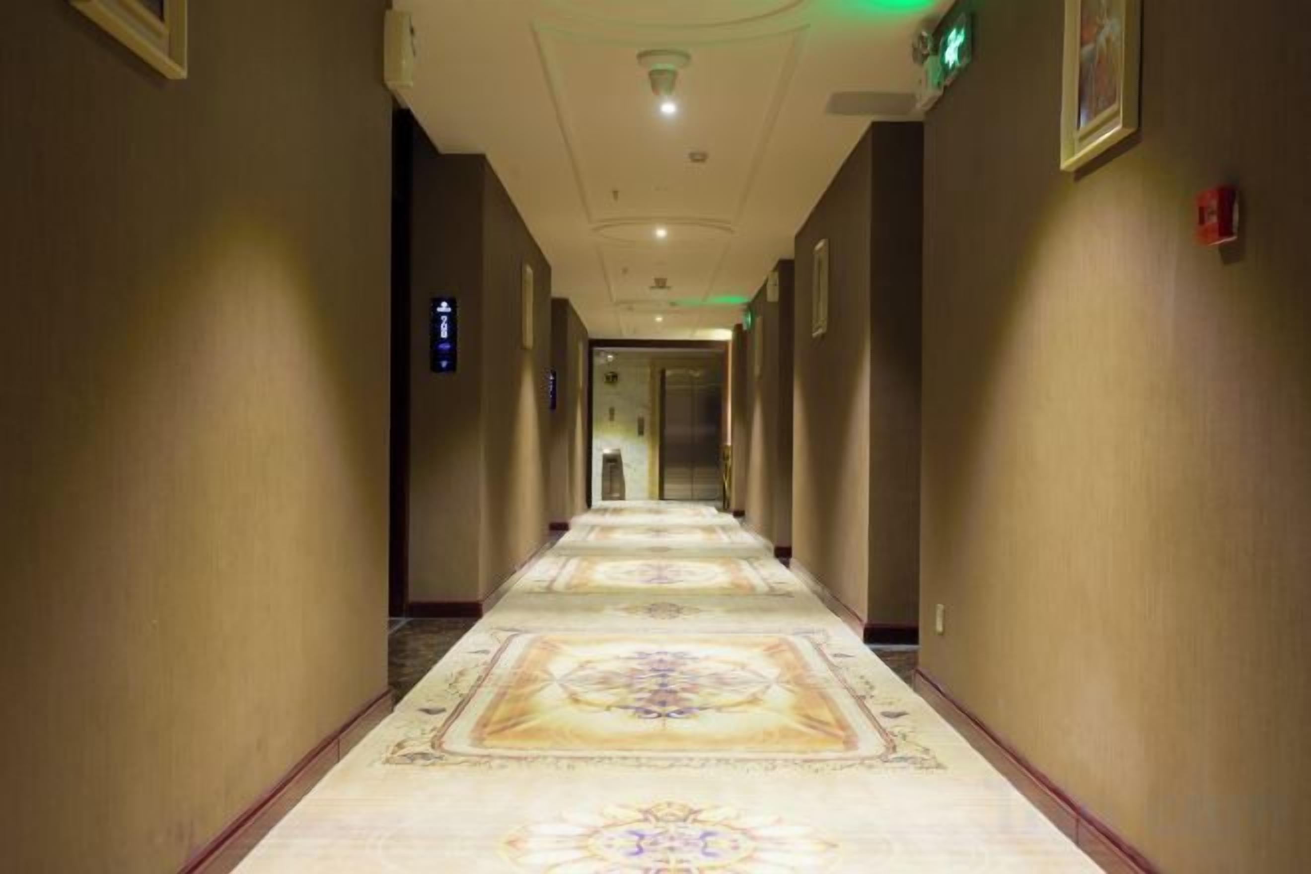 A hotel corridor with a patterned carpet, a fire alarm on the wall, and a door at the end.