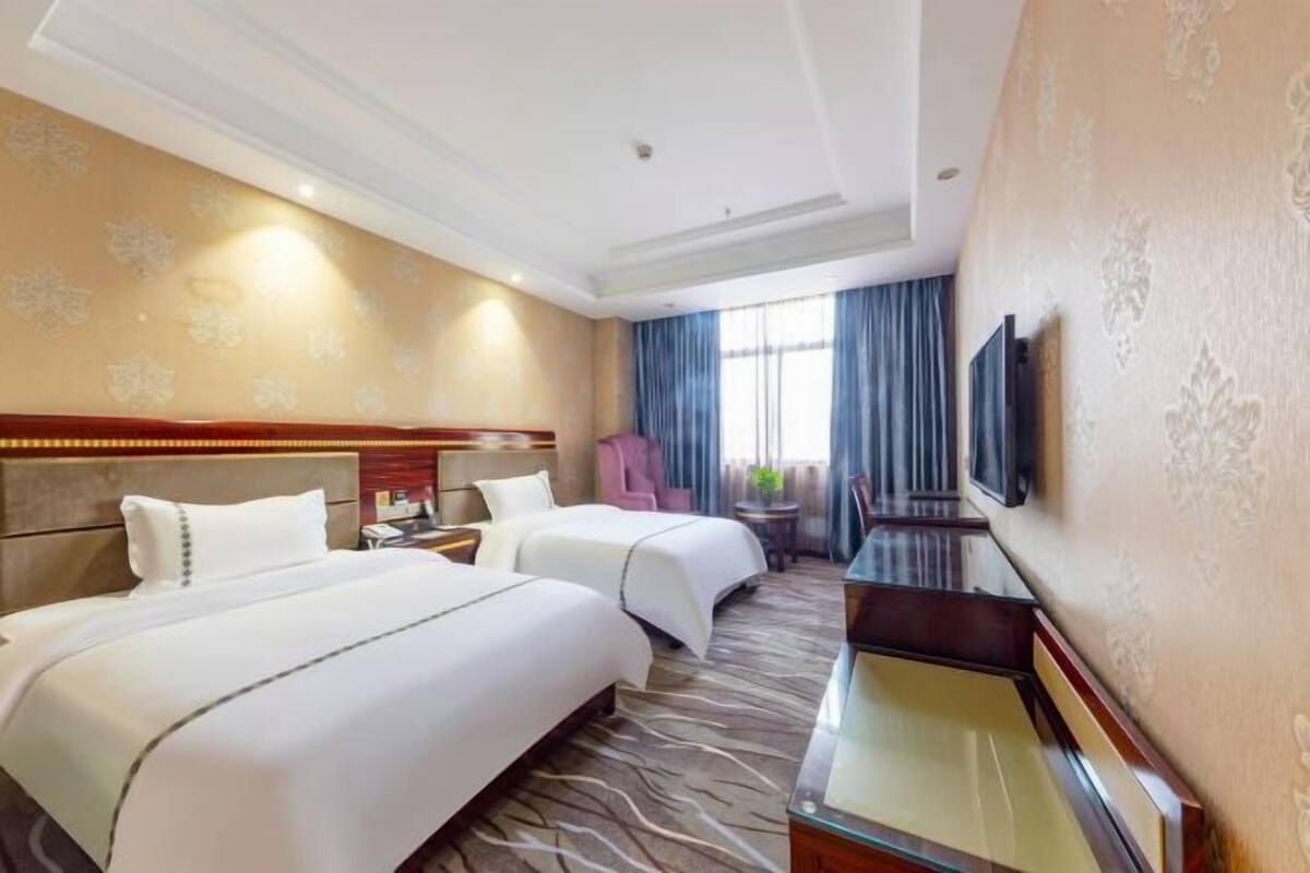 A hotel room with two beds, a desk, a TV, and a window with curtains.