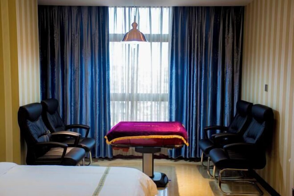 A hotel room with a bed, two black chairs, a massage table, and a large window with blue curtains.