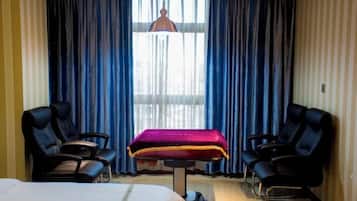 Una camera d'albergo con un letto, due sedie nere, un lettino da massaggio e un'ampia finestra con tende blu.