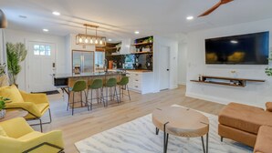 A modern living room with a large TV, yellow chairs, a wooden coffee table, and a kitchen area with a bar and stools. - Luxe 4-Bed Retreat w/ Game Room & Deck – 10 Min to Downtown (Nashville)