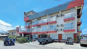 A multi-story hotel with a red and white facade, balconies, and a parking area with vehicles.