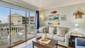 A spacious living room with a large sliding glass door leading to a balcony, a grey sofa, and a small table with a decorative item. - EndlessWaves Oceanfront Gulf View Resort 2 Pools (Galveston)