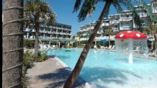 A pool area with palm trees, a water slide, and a building with balconies in the background. - EndlessWaves Oceanfront Gulf View Resort 2 Pools (Galveston)
