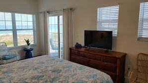 A bedroom with a large bed, a dresser, a television, and a view of the beach through the window.