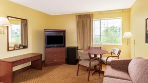 Camera d'albergo con una scrivania in legno, televisione, tavolo da pranzo con sedie, divano e un'ampia finestra con tende.