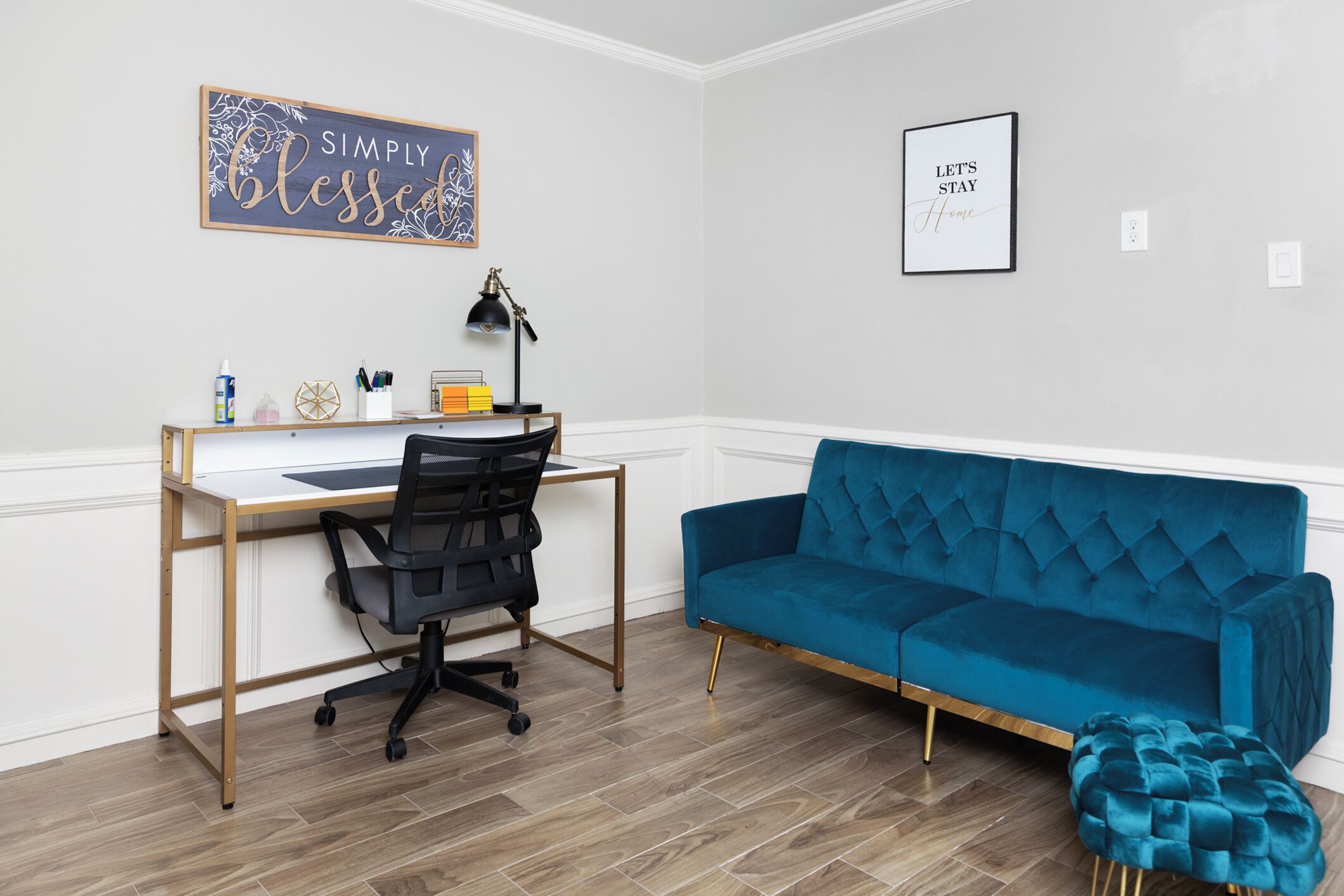 Une pièce comprenant un bureau en bois, un fauteuil de bureau noir, un canapé en velours bleu et un petit pouf. Deux panneaux encadrés sont accrochés au mur et une lampe se trouve sur le bureau.