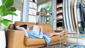 Una persona estesa en un sofà marró llegint un llibre en una habitació amb una porta de vidre que dona a l'exterior, un suport per a taules de surf i una planta en test.