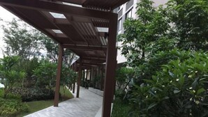 Un paseo techado con piso de baldosas, rodeado de exuberante vegetación y un edificio al fondo.