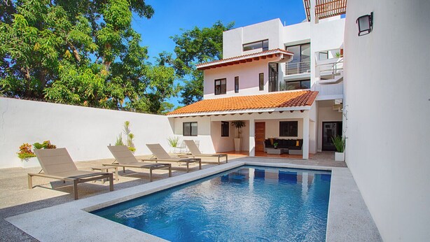 A swimming pool with lounge chairs, a white building with a red-tiled roof, and a balcony.