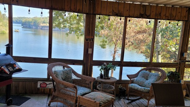 A covered outdoor deck with wicker furniture, a ceiling fan, and a view of a lake.