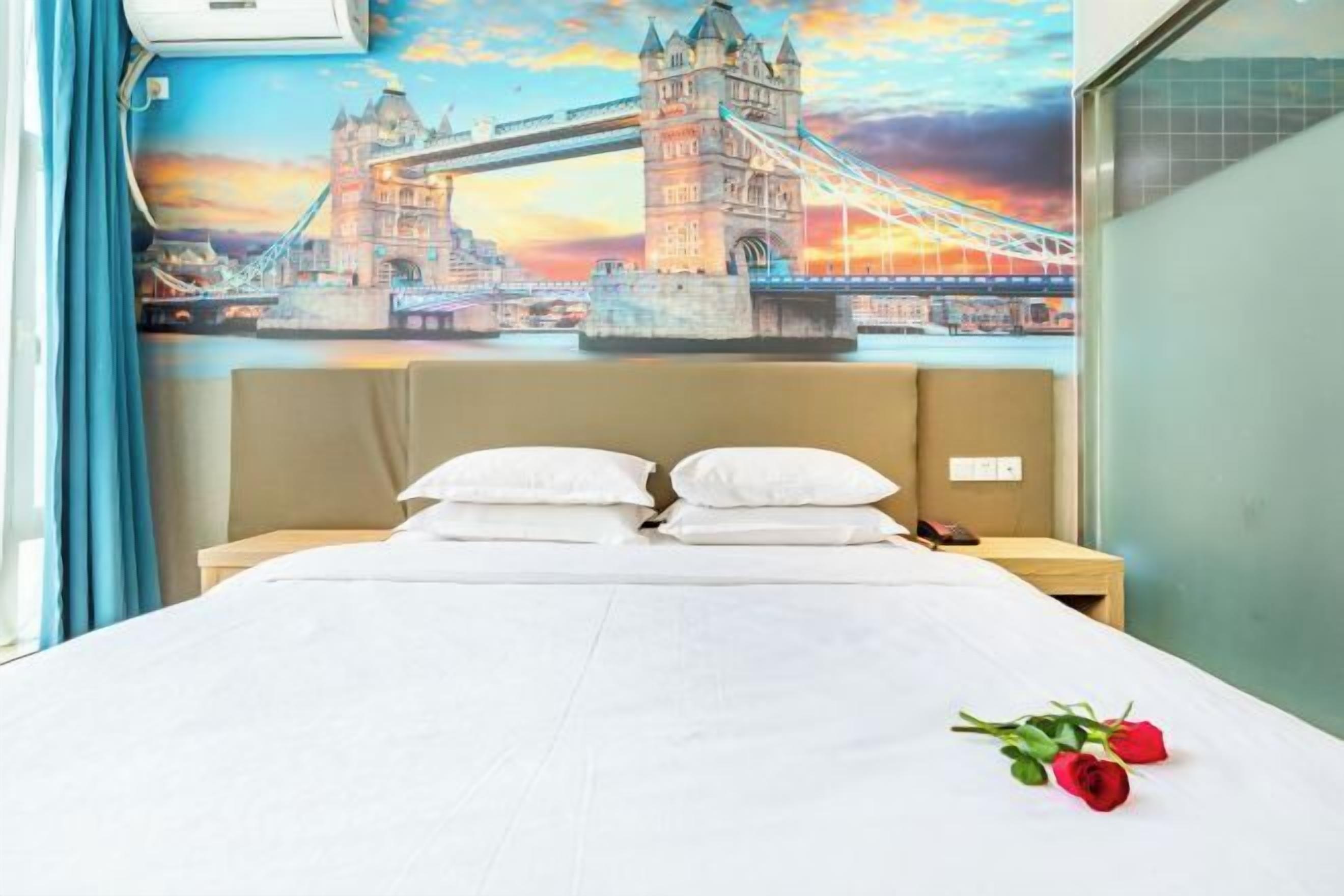 Ein Hotelzimmer mit Bett, Kissen, einem Nachttisch, einer Wand mit einem Wandgemälde einer Brücke und einem Fenster mit Vorhängen.