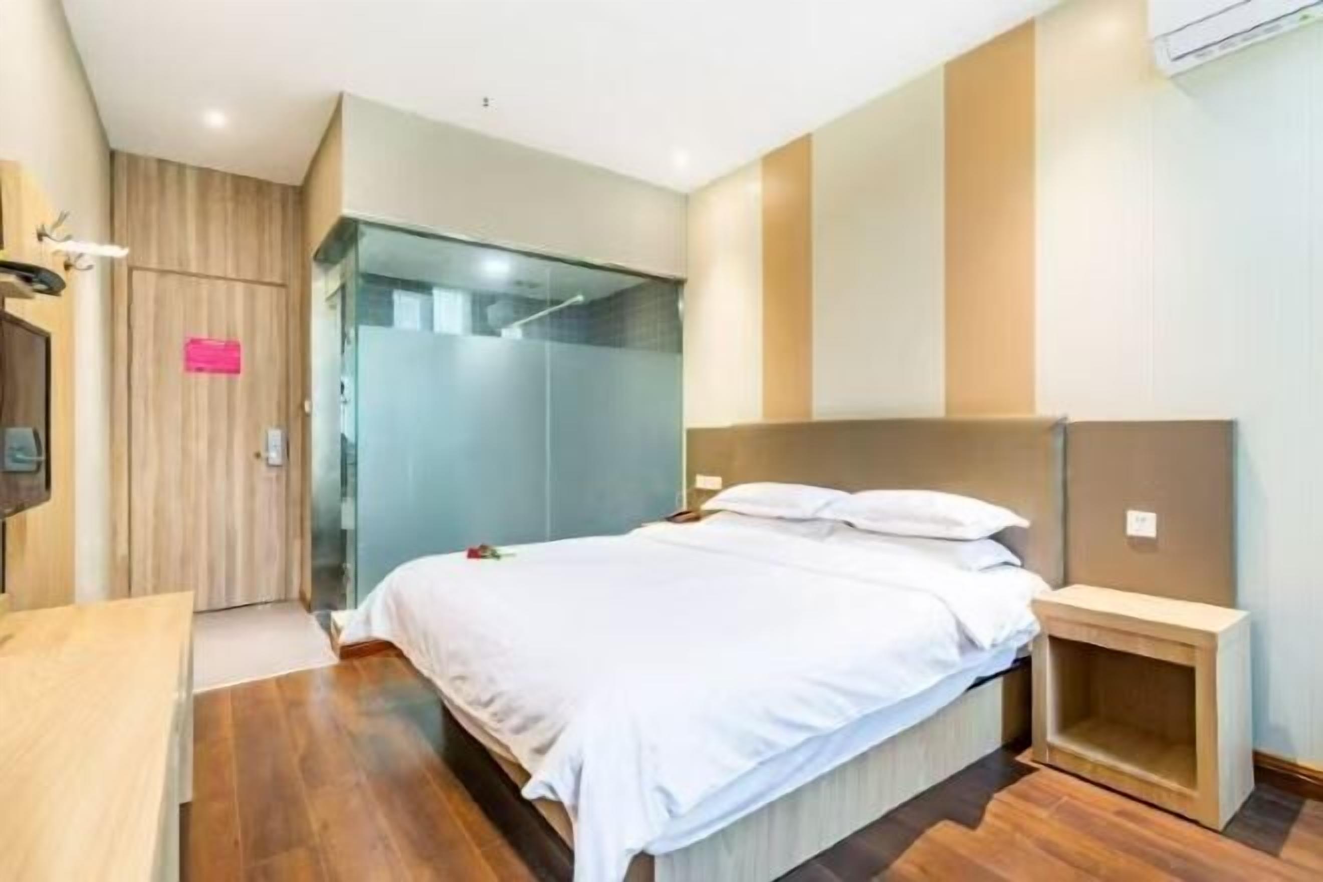 A hotel room with a bed, a wooden wardrobe, a glass shower enclosure, and a small bedside table.