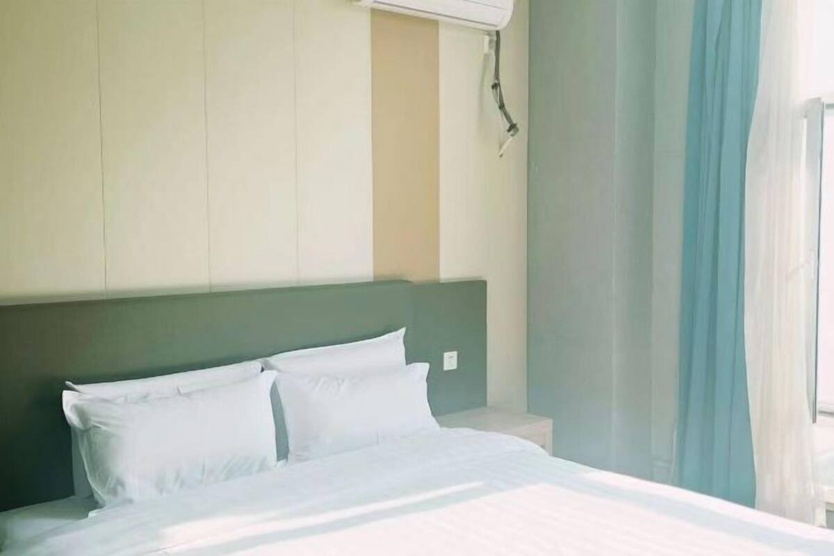 Una habitación de hotel con una cama, cabecera, aire acondicionado mural y cortinas traslúcidas.