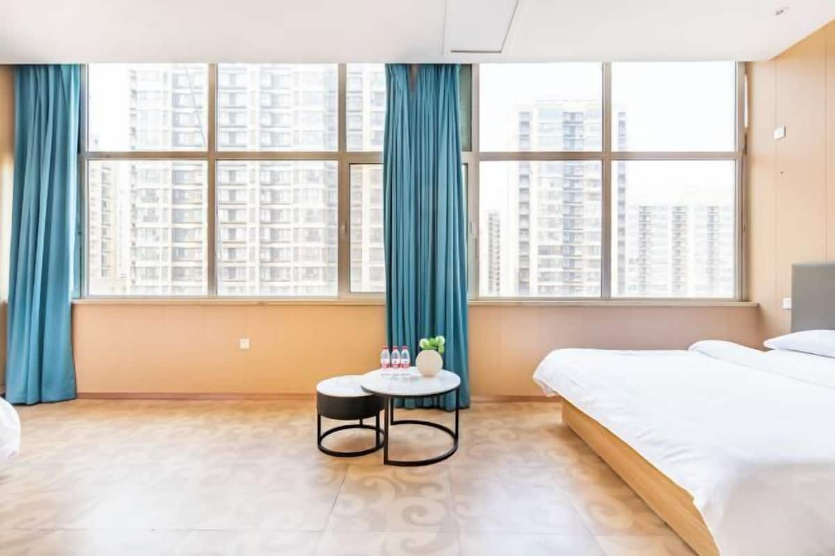 Habitación de hotel con cama, una pequeña mesa redonda con una planta, ventana con cortinas y vistas a edificios altos.