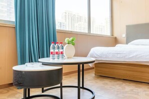 A hotel room with a bed, a round table with a vase and water bottles, a round ottoman, and a view of buildings through a window. - City 118 Chain Inn Qingdao Zhengyang Road Liqun (Qingdao)