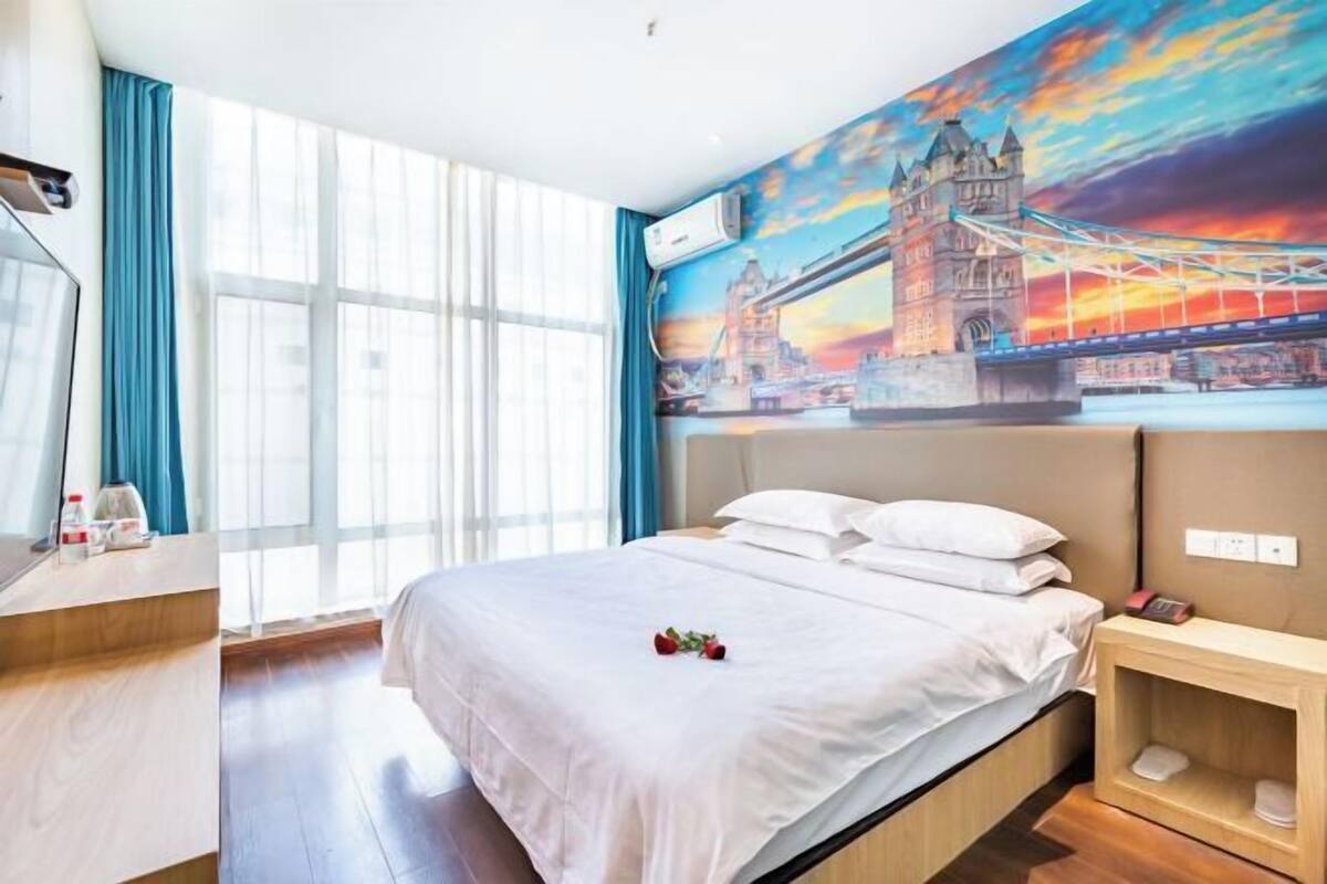 Habitación de hotel con cama, escritorio, silla, ventana con cortinas y un mural en la pared de un puente.