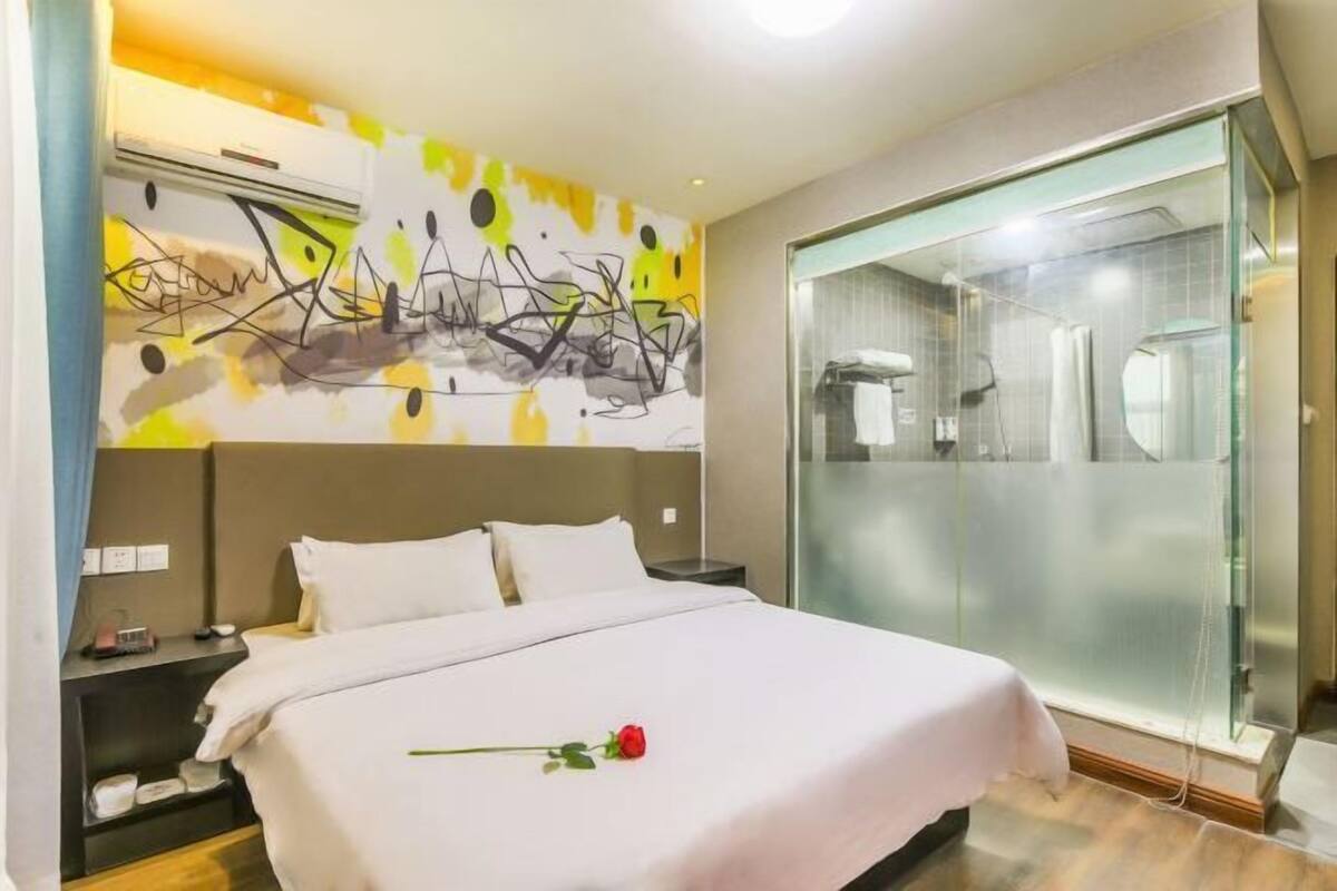 Una habitación de hotel con cama, mesita de noche, una obra de arte en la pared y un baño con puerta de cristal.