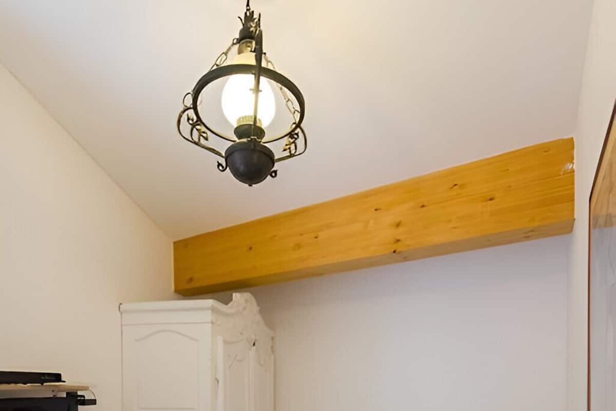 Um quarto com teto de madeira, uma cama com colcha colorida, um guarda-roupa branco, uma mesa de cabeceira com abajur e uma televisão sobre um suporte.