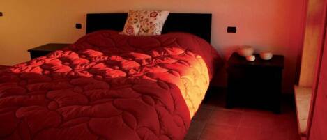 A bedroom with a red bedspread, a nightstand with a lamp, and a pillow with a floral pattern.