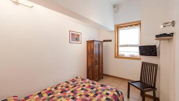 A bedroom with a bed, a wooden wardrobe, a chair, a TV mounted on the wall, and a window with curtains.