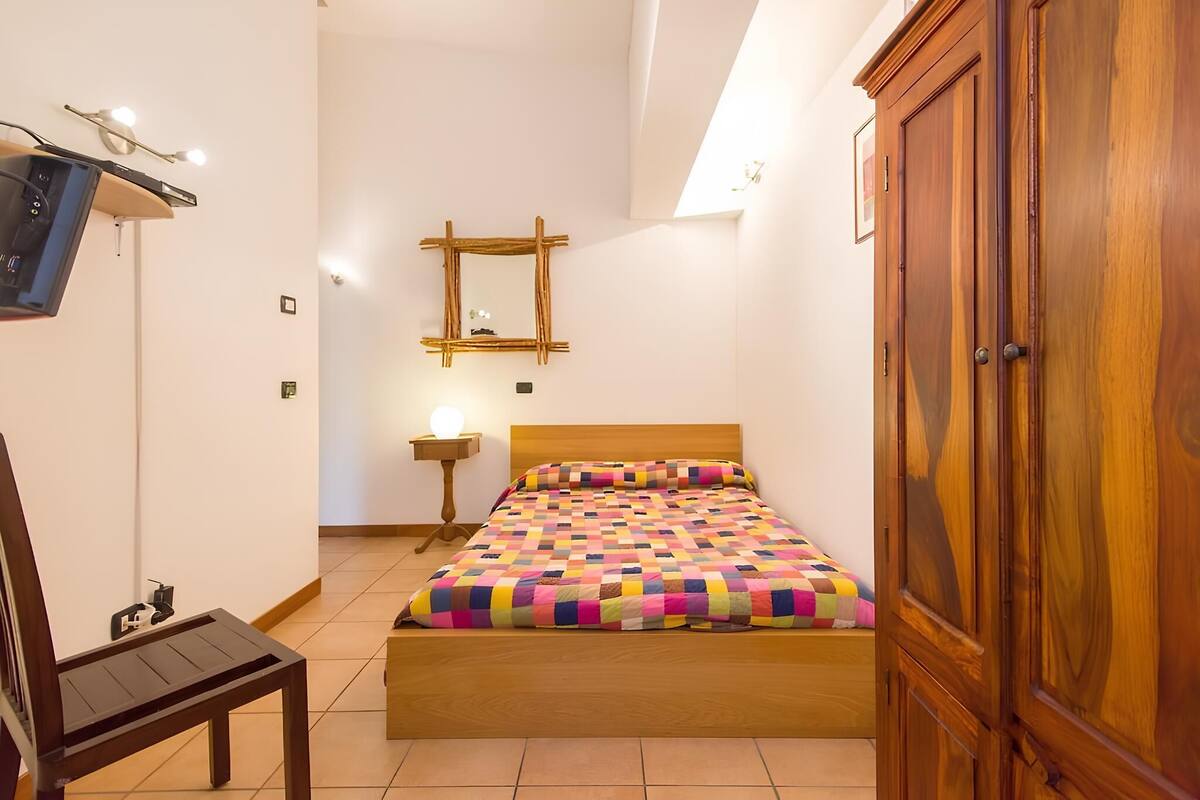 Quarto com cama de madeira, guarda-roupa de madeira, mesa de cabeceira com luminária e espelho fixado na parede.