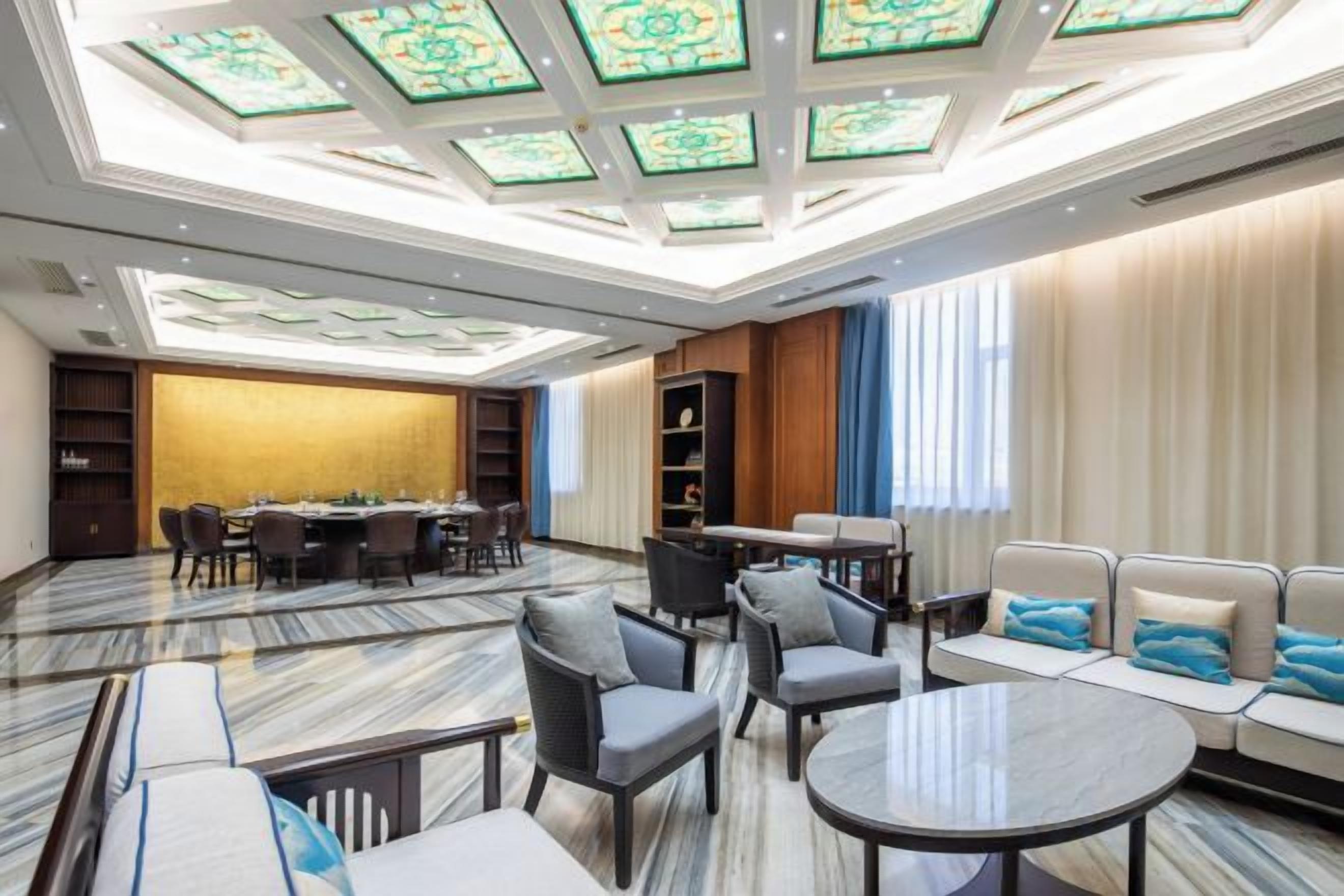 Un hall d’hôtel avec un plafond haut, un espace salon avec des canapés et des fauteuils, un coin repas avec des tables, et une grande fenêtre avec des rideaux.