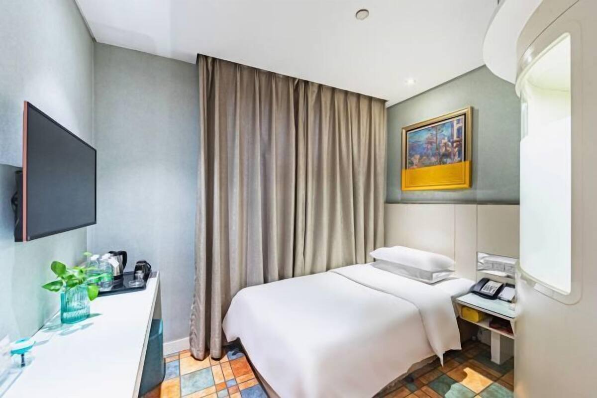 Ein Hotelzimmer mit Bett, Schreibtisch mit Stuhl, Wandfernseher, Vorhängen an einem Fenster und einem Wandbild.