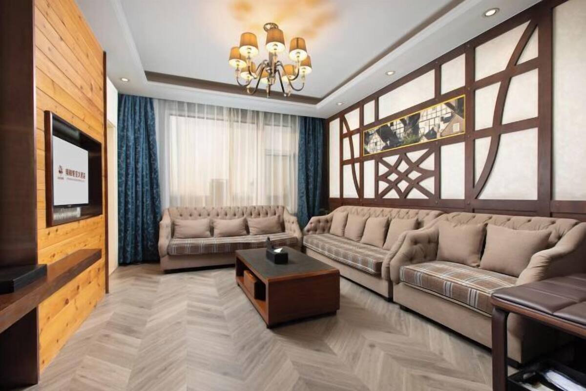 Ein Wohnzimmer mit Fischgrätparkett, zwei Sofas, einem Couchtisch, einem an einer Holzwand montierten Fernseher und einem großen Fenster mit Vorhängen.
