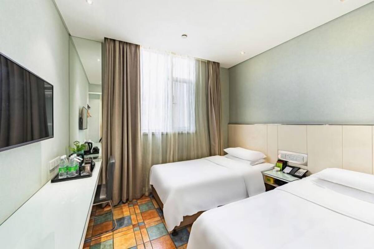 Ein Hotelzimmer mit zwei Betten, einem Schreibtisch mit Stuhl, einem Fernseher und einem Fenster mit Vorhängen.