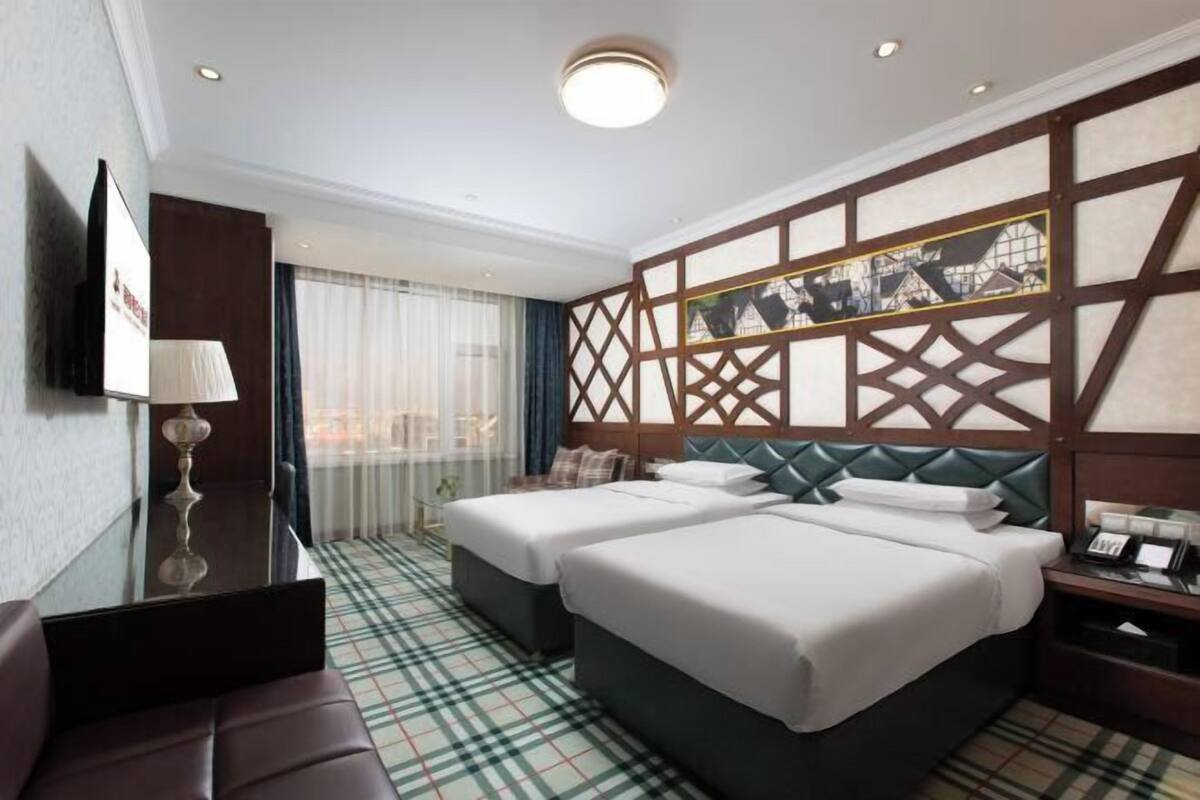 Ein Hotelzimmer mit zwei Betten, einem Fernseher, einem Schreibtisch, einer Lampe und Blick auf Gebäude durch eine Wand mit Muster.