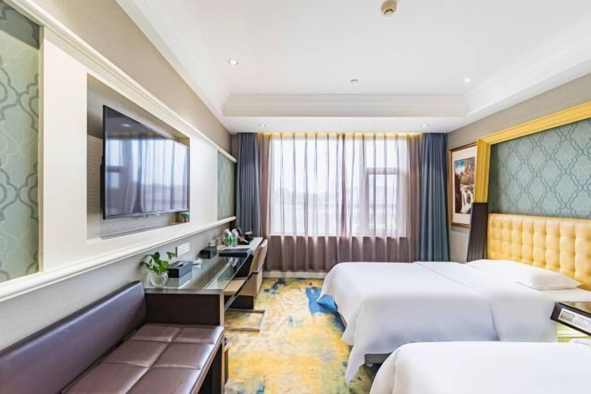Ein Hotelzimmer mit zwei Betten, einem Fernseher, einem Schreibtisch mit Stuhl, einer Bank und einem großen Fenster mit Vorhängen.
