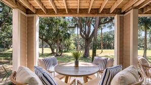 A covered outdoor seating area with wicker chairs and a round table, surrounded by trees. - Luxury Villa, Club Access, Beach, Pet-Friendly (Seabrook Island)
