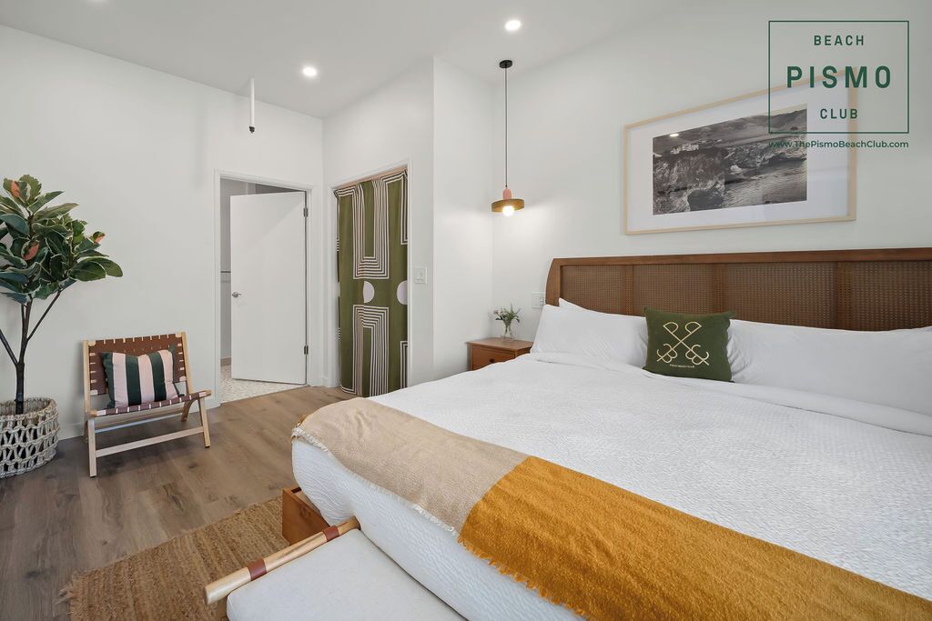 Quarto de hotel com uma cama grande, cabeceira de madeira, mesa de cabeceira, um banquinho, uma planta em vaso e um quadro na parede.