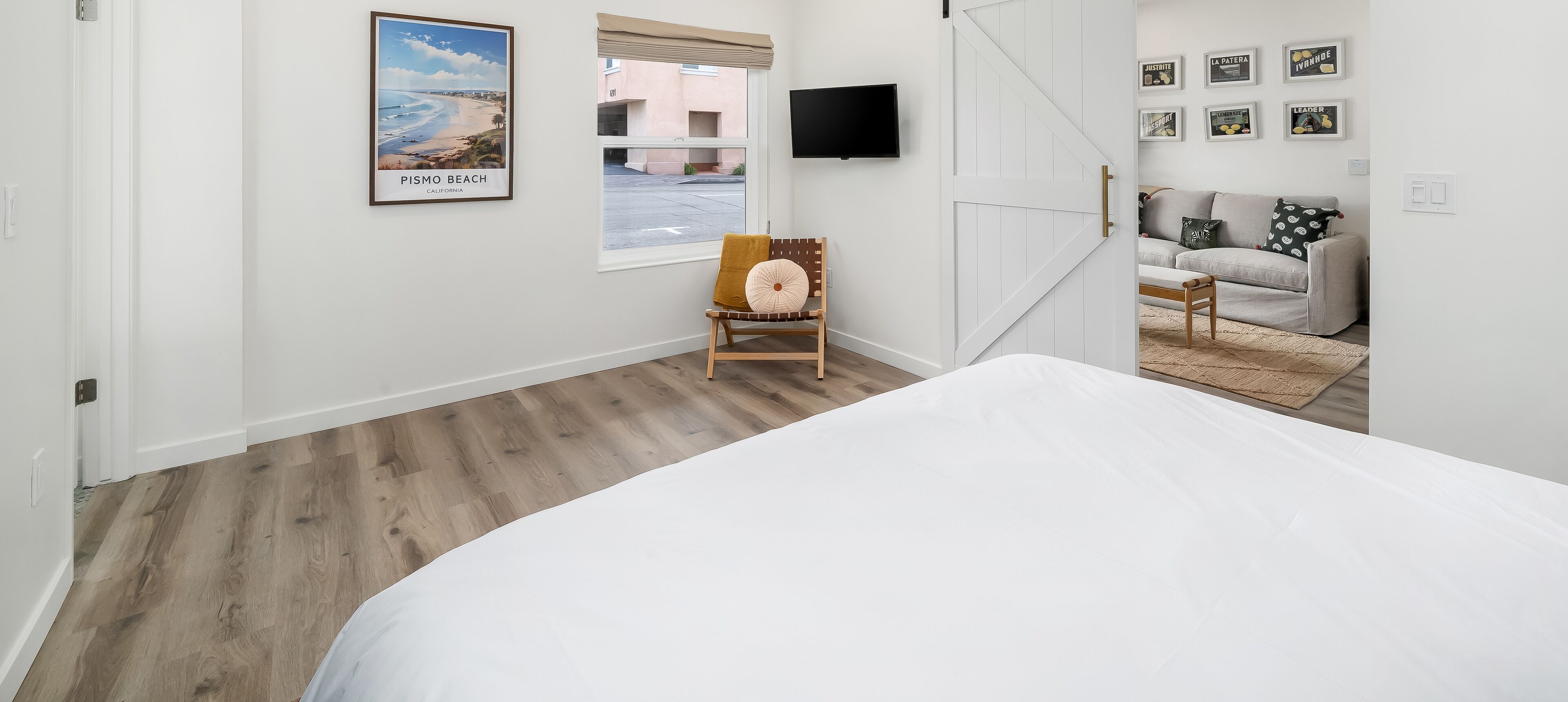 A bedroom with a large bed, a chair with a cushion, a TV mounted on the wall, and a window with a view of the outside.