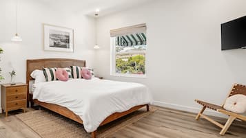 Ein Schlafzimmer mit einem hölzernen Bett, einem Nachttisch, einem Beistelltisch, einem Sessel, einem Fenster mit Aussicht und einem Wand-Fernseher.