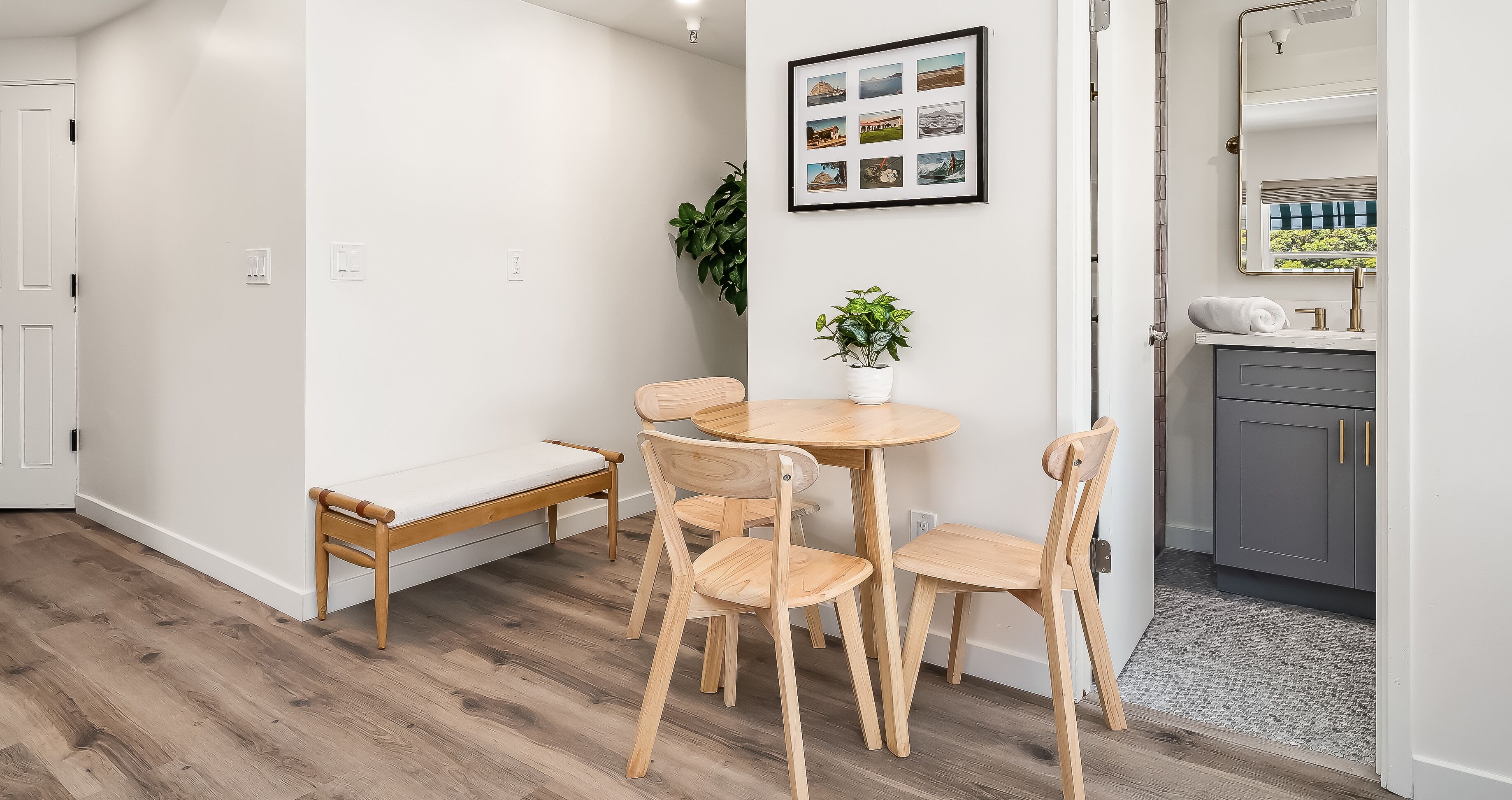 A modern, minimalist room with a wooden dining table and chairs, a bench, a potted plant, and a framed photo collage on the wall.