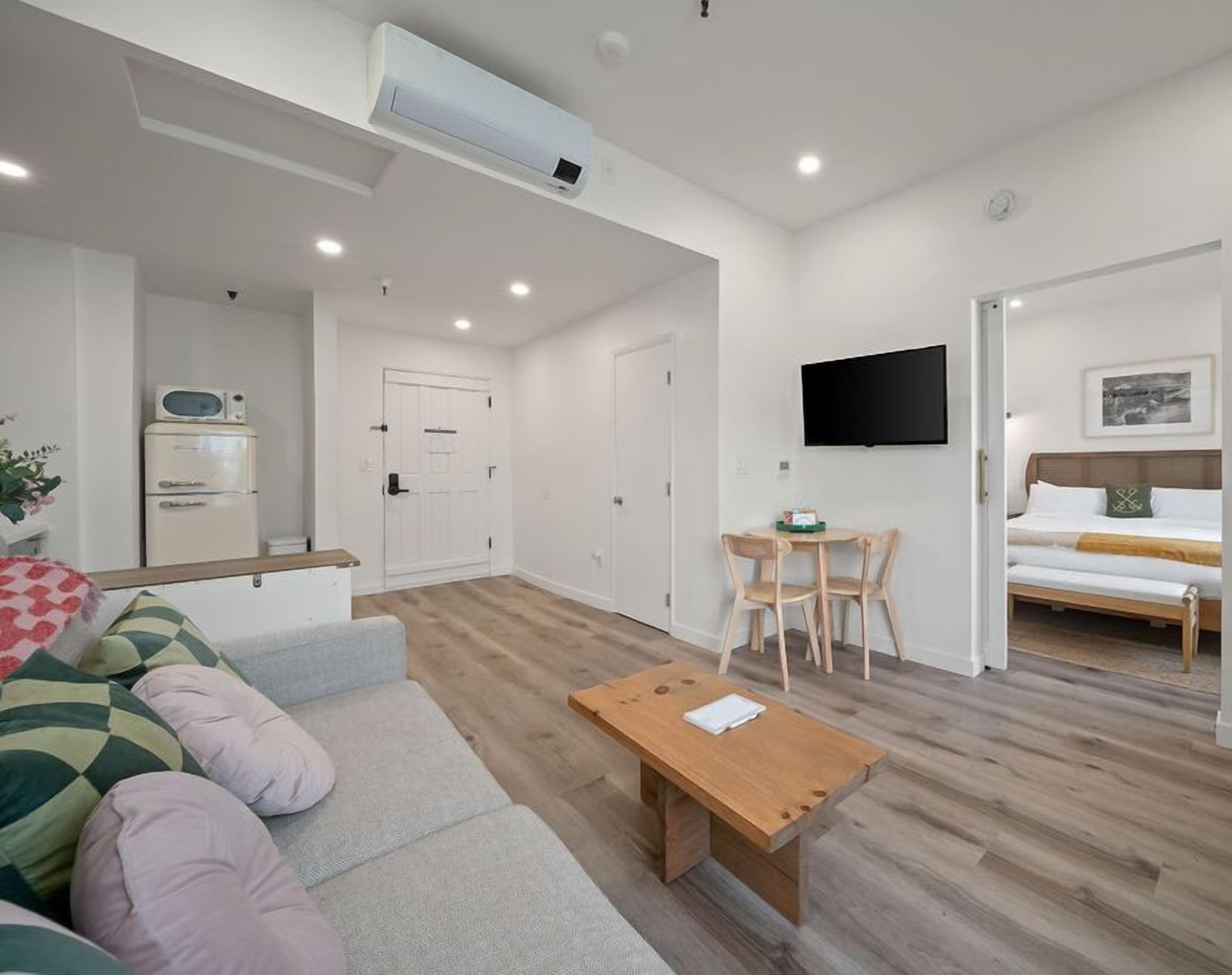 Ein modernes Wohnzimmer mit einer Couch, einem Holztisch, einem an der Wand montierten Fernseher und einem Bett im angrenzenden Raum.