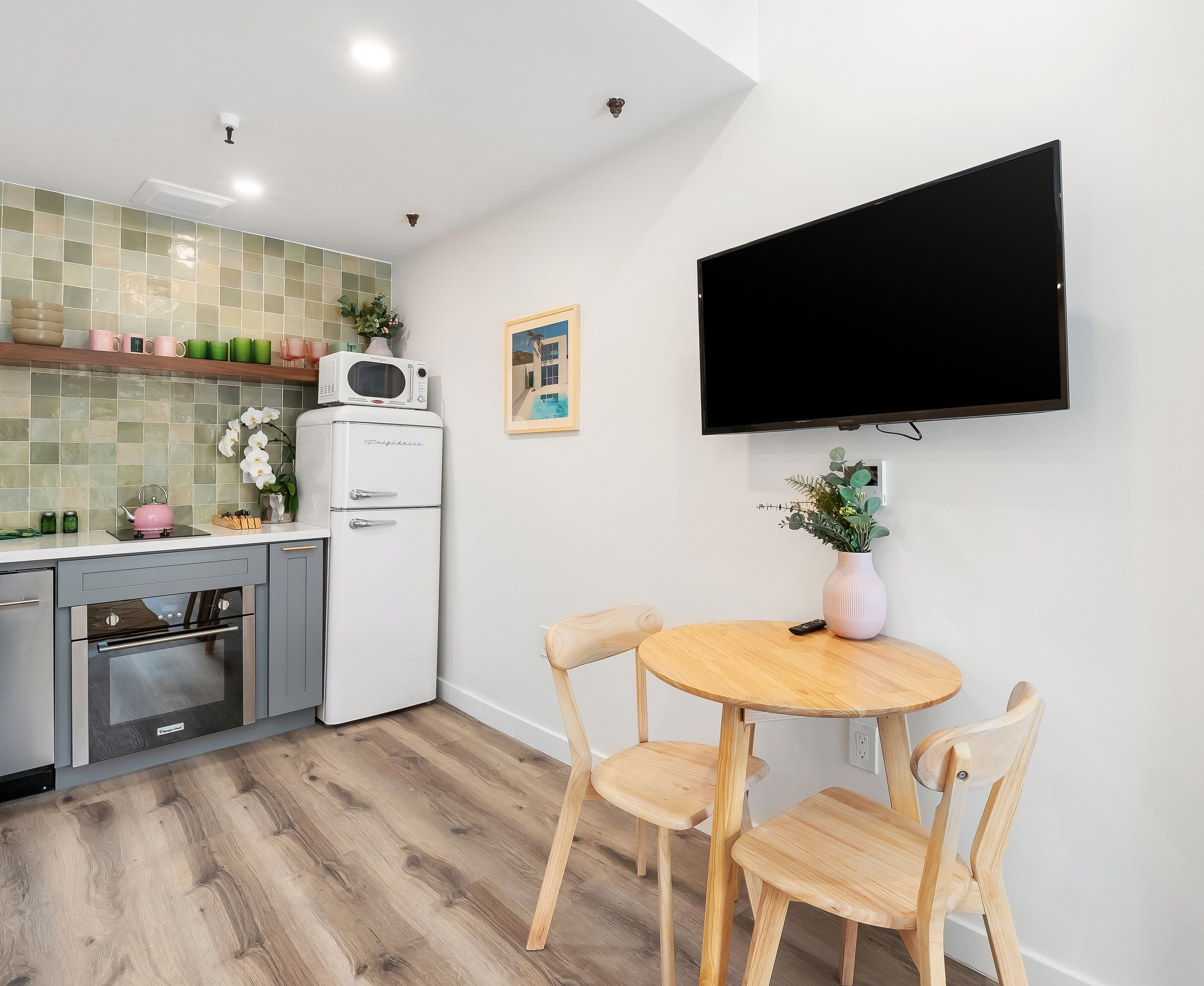 A modern kitchen with a microwave, refrigerator, and oven. There is a dining table with chairs and a TV mounted on the wall.