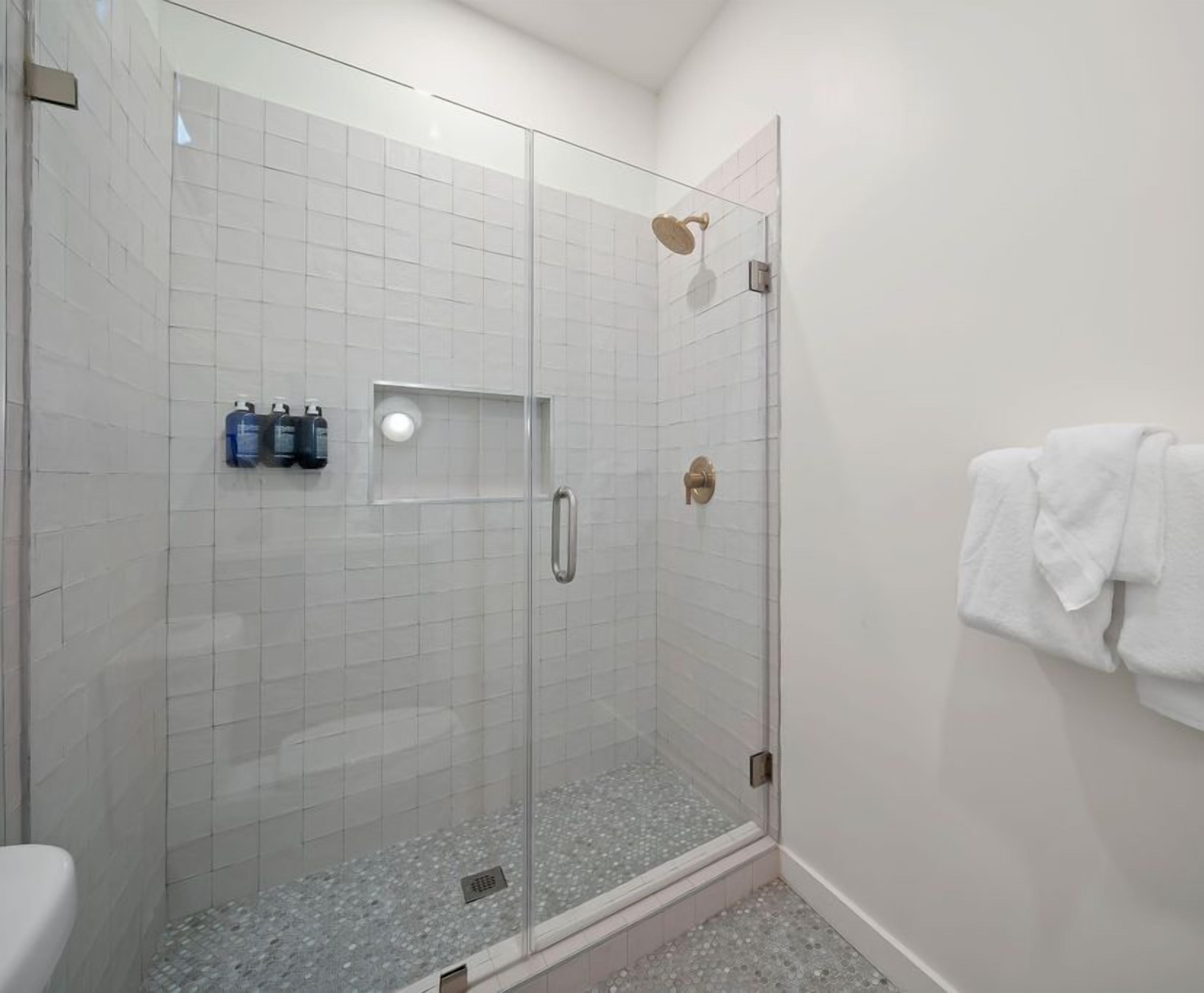 A glass-enclosed shower with a tiled floor, a towel rack with a white towel, and two bottles of shampoo on the wall.