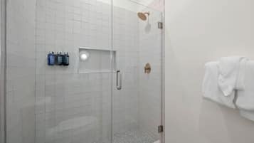 A glass-enclosed shower with a tiled floor, a towel rack with a white towel, and two bottles of shampoo on the wall.