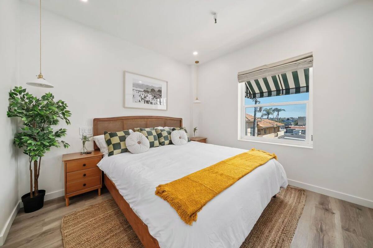 Un dormitorio con cama, mesita de noche, planta en maceta, ventana con vista y un cuadro en la pared.