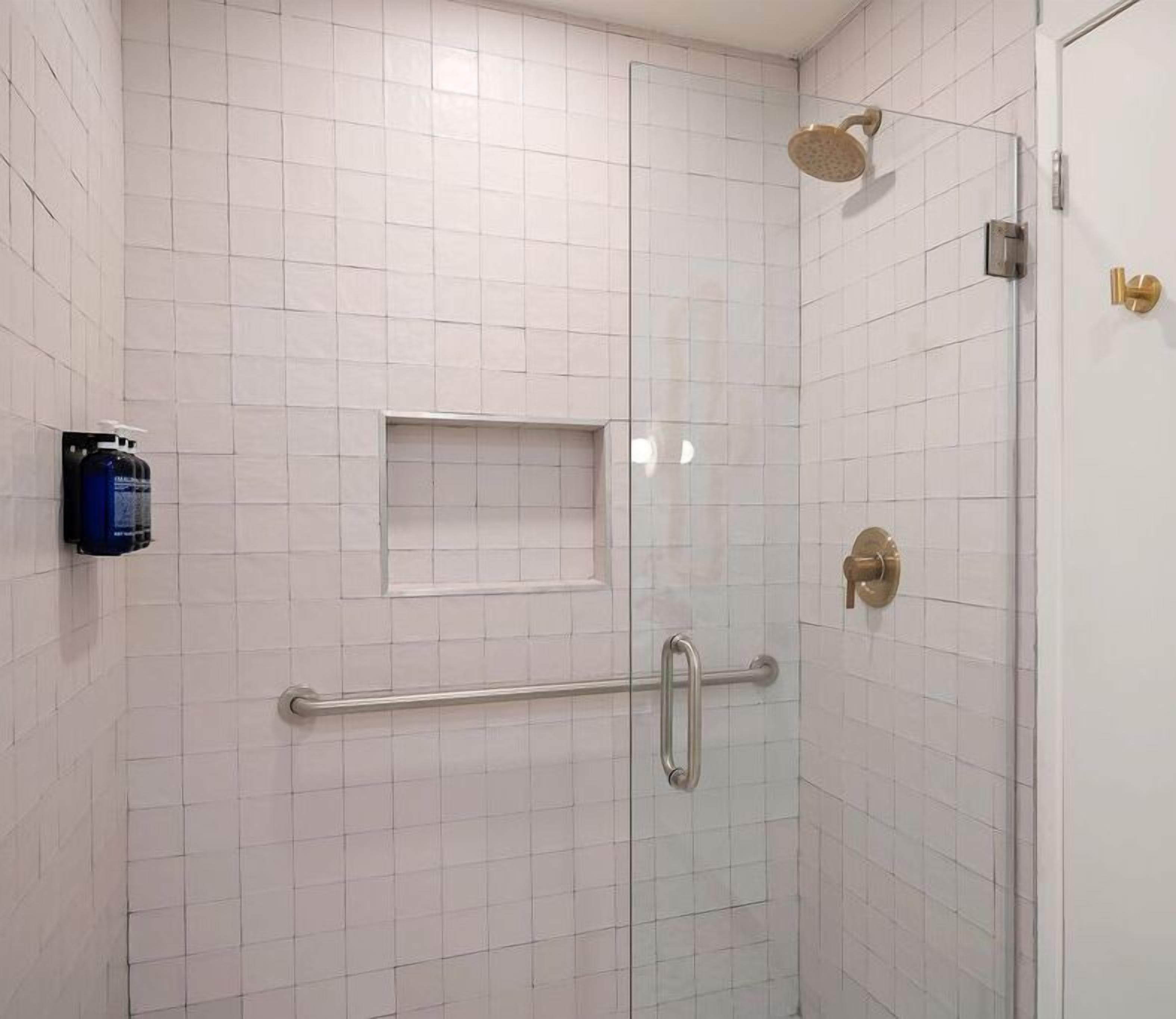 A shower area with white tiled walls, a glass door, a handheld showerhead, a grab bar, and a soap dispenser.